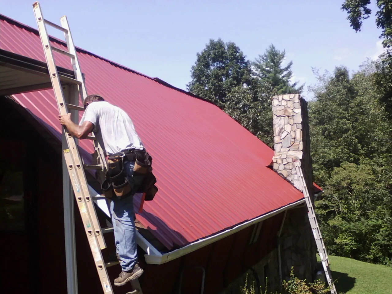 Roofer on a ladder beside a red metal roof and stone chimney — Soffit & Fascia Repair by Roofing Brothers in Bel Air